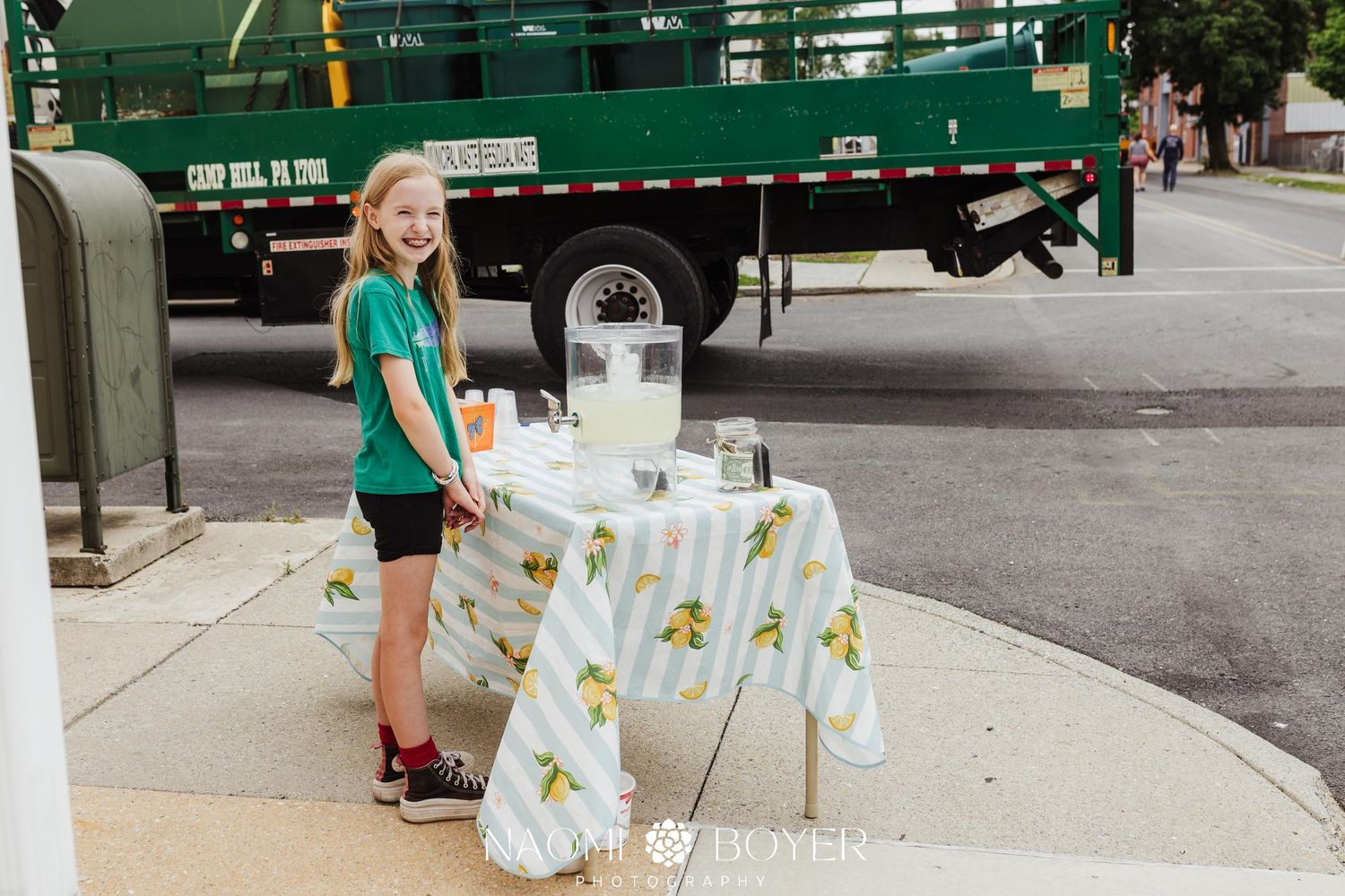Young girl smiling at a lemonade stand on a sidewalk with a green truck in the background.
