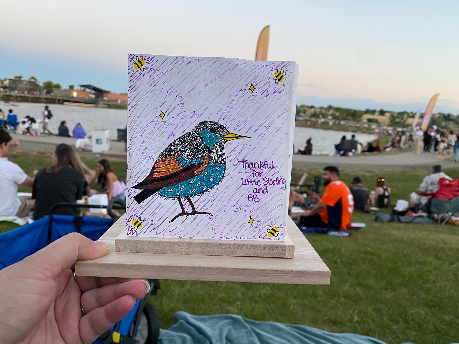 Hand-held colorful bird artwork on canvas with bees, displayed outdoors at a community event on a sunny evening.