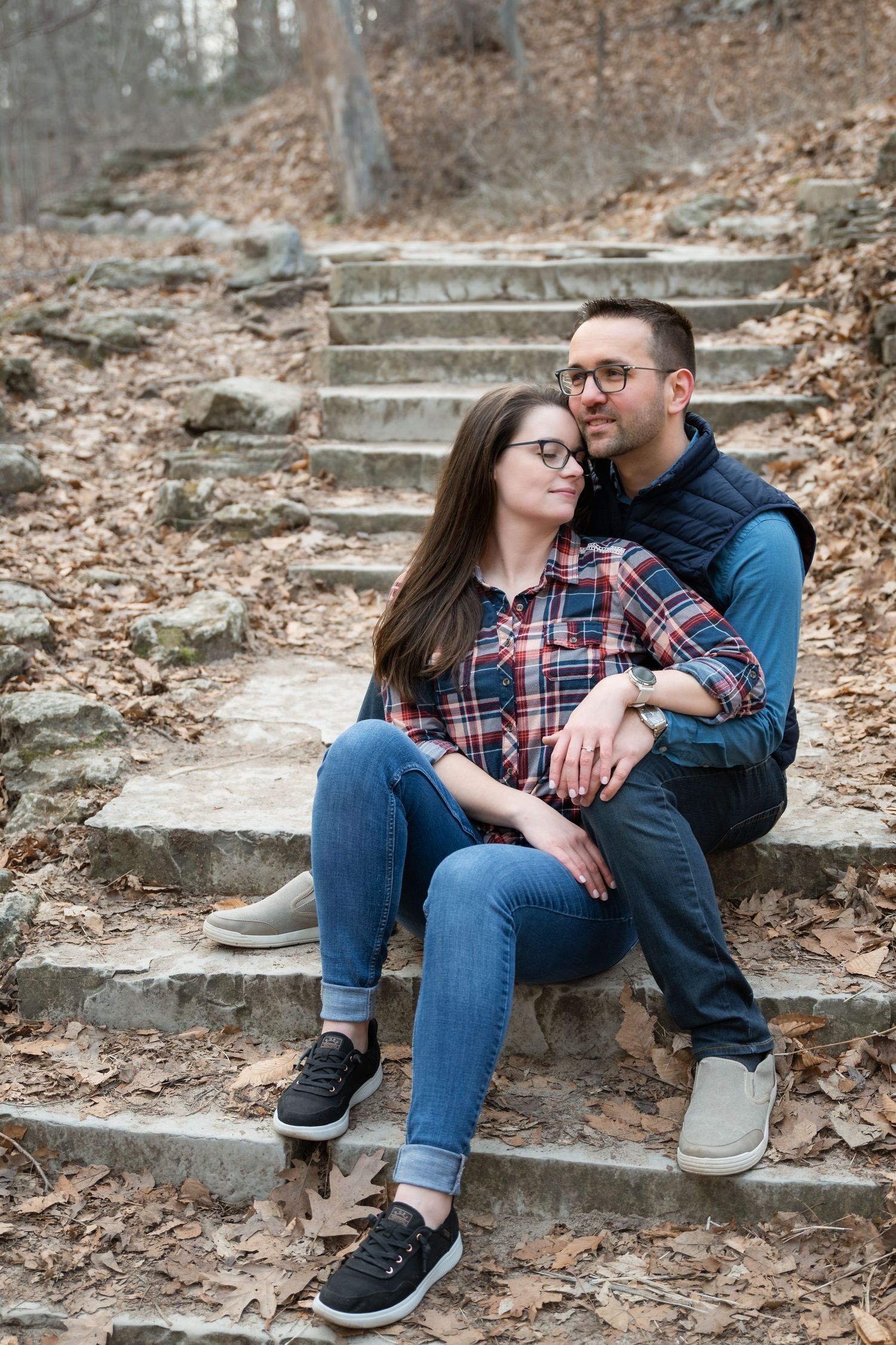 Couple smiling at each other leaning against a tree in a forest, wearing jeans and casual flannel shirts.