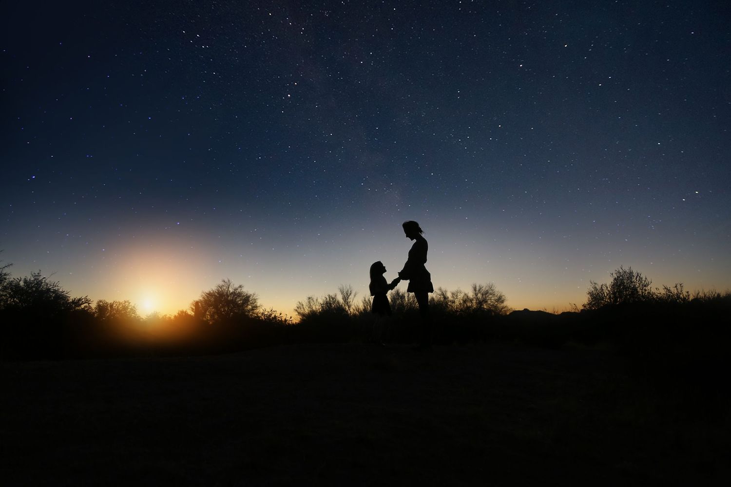 Silhouettes of a mother and child stand facing each other under a starry night sky with a glowing sunset on the horizon.