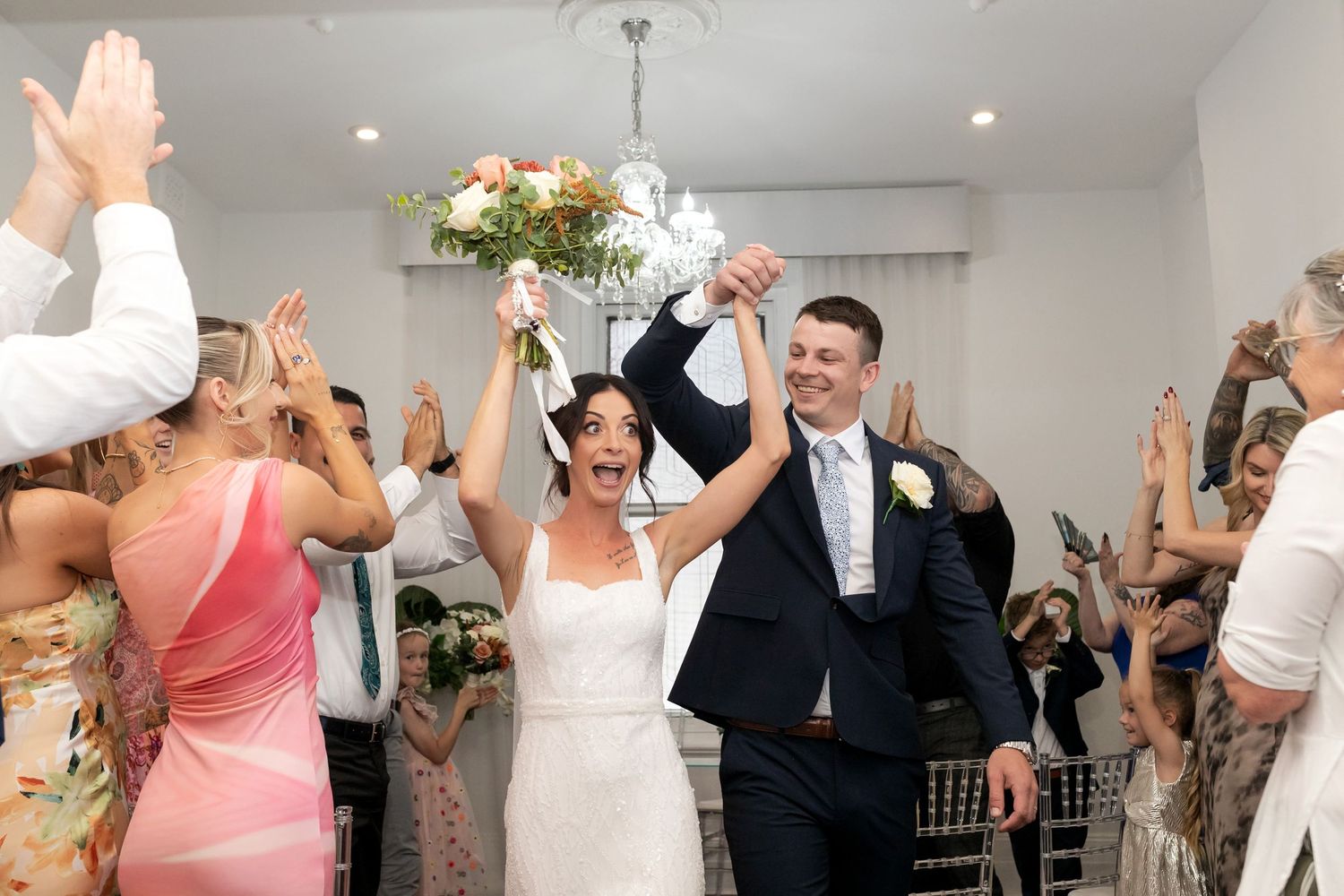 Joyful newlyweds celebrate their wedding ceremony exit with raised hands, surrounded by cheering guests in a bright venue.