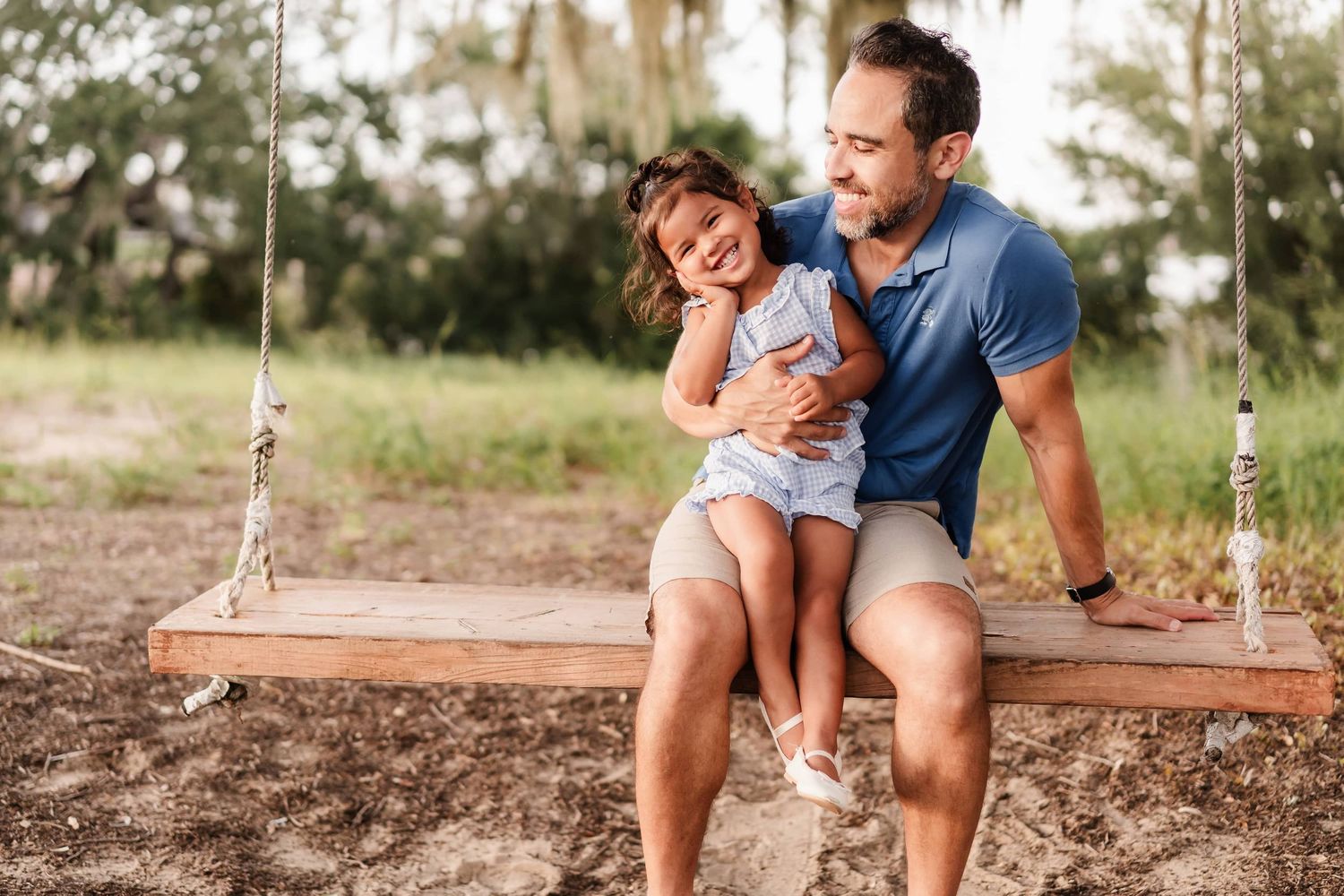 Parent in a red shirt lifts a giggling child wearing a red outfit up in the air at sunset in a grassy field.