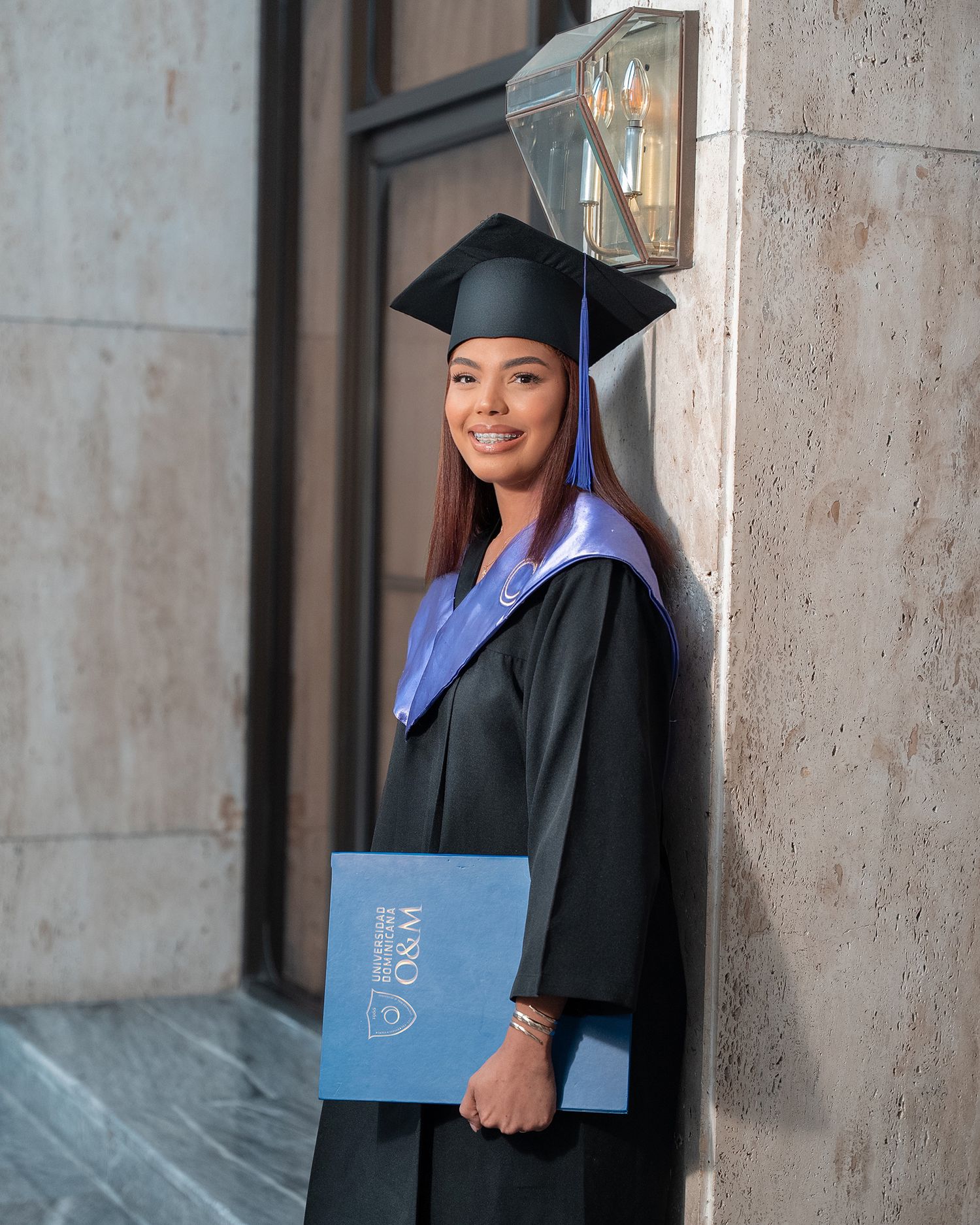 Smiling graduate in black cap and gown sits holding diploma folder, celebrating academic achievement.