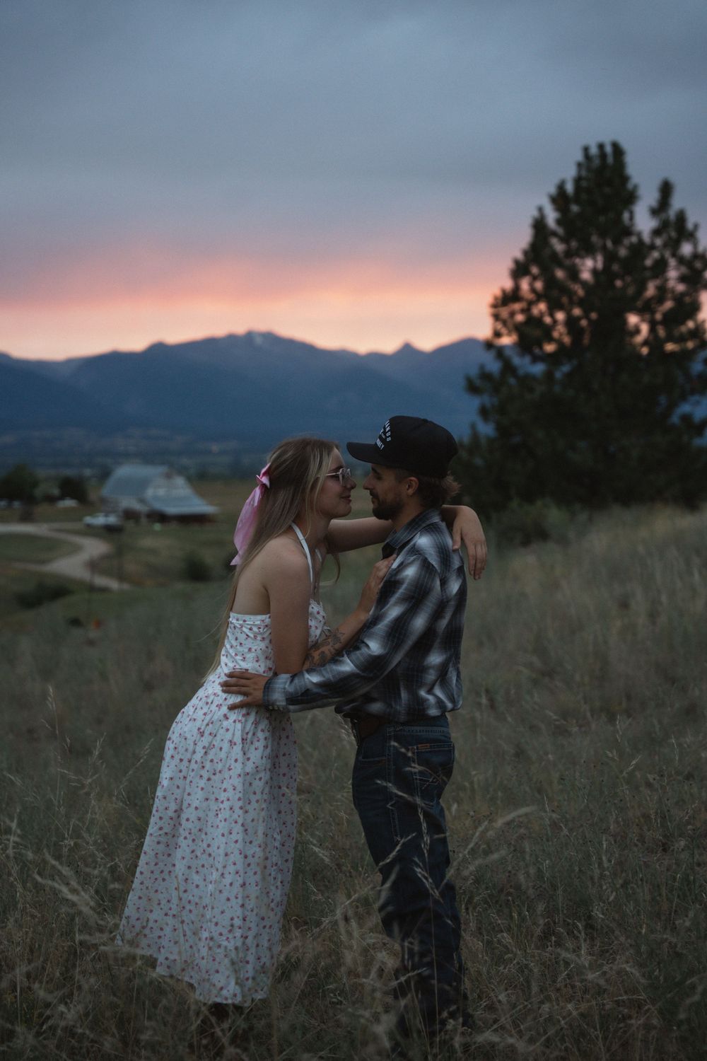 Couple embracing in a field at sunset with mountain backdrop, woman in floral dress and man in plaid shirt and cowboy hat.
