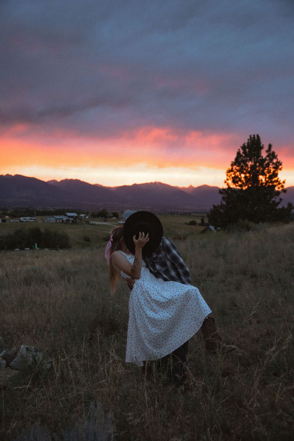 Couple sharing a romantic kiss in a field at sunset with vibrant pink and purple sky and mountain backdrop.