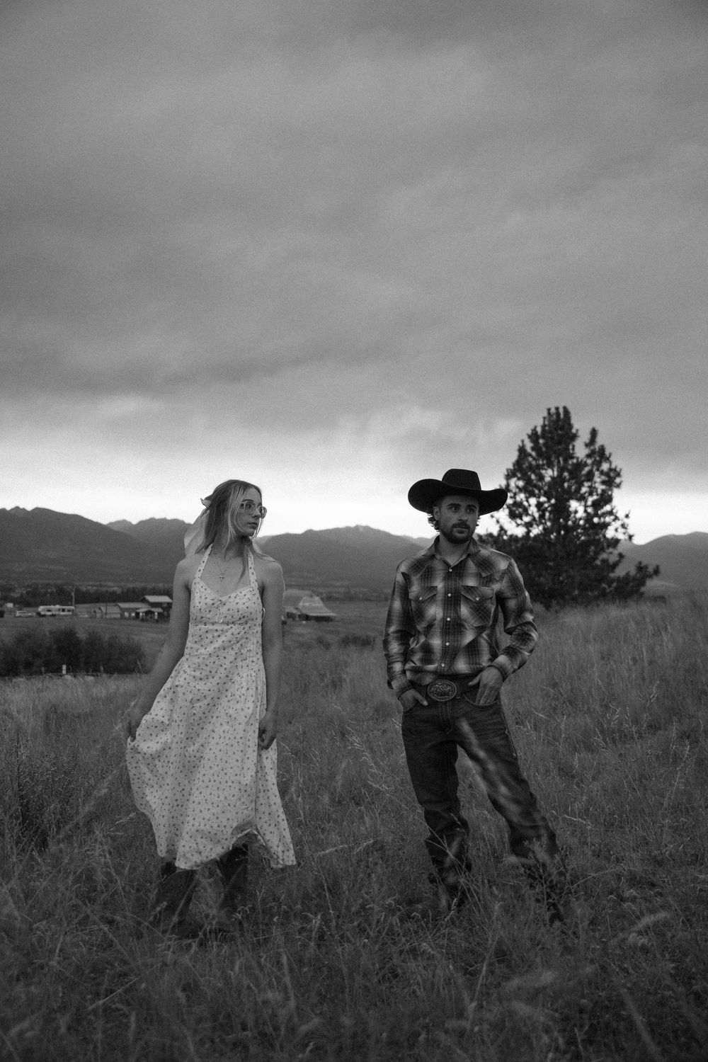 Black and white photo of a couple standing in a field with mountains and dramatic cloudy sky in the background.