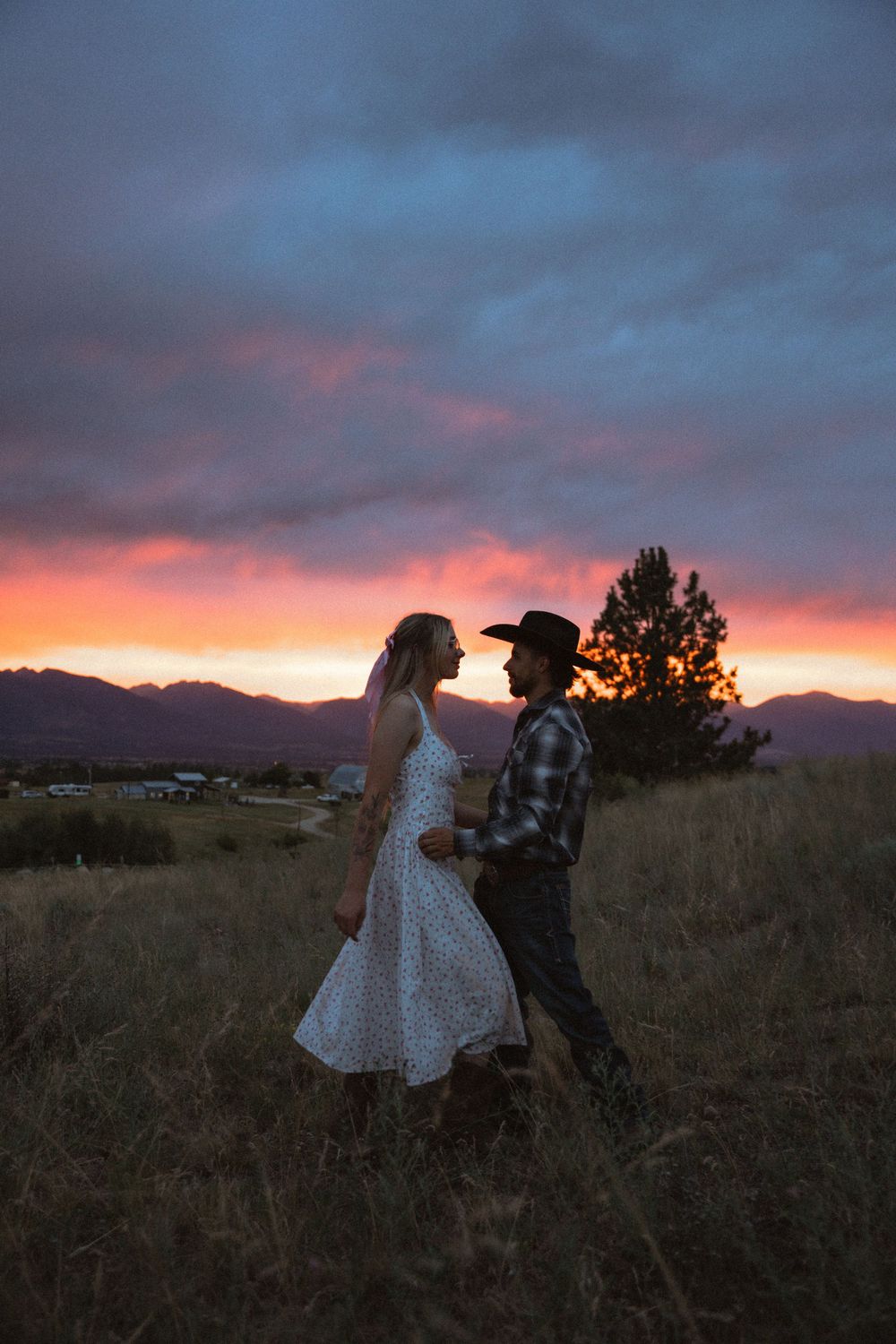 Couple dancing in a field at sunset with vibrant pink and blue sky, mountains, and trees in the background.