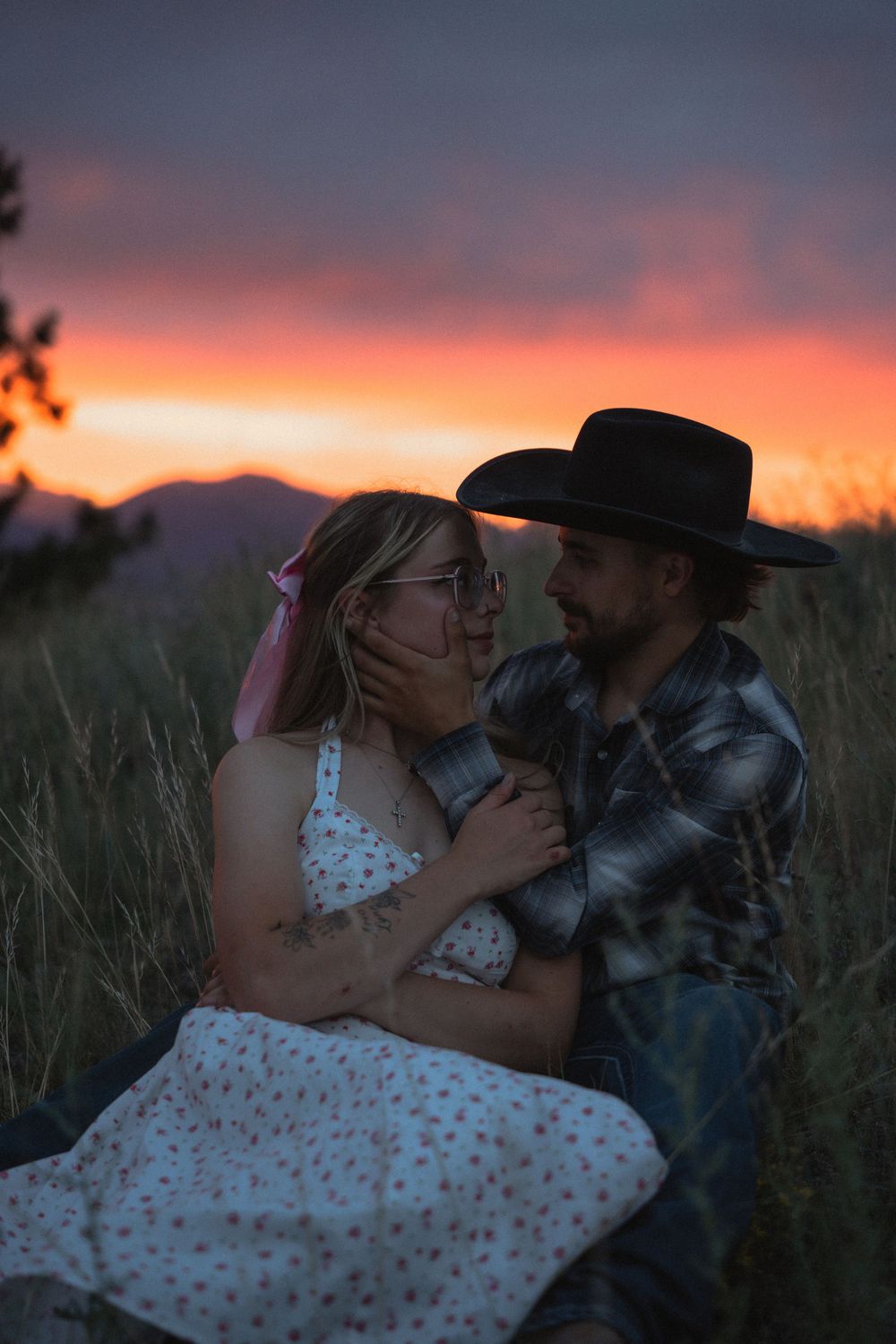 Couple sharing a romantic moment in tall grass at sunset, man in black cowboy hat and woman in floral dress embracing.