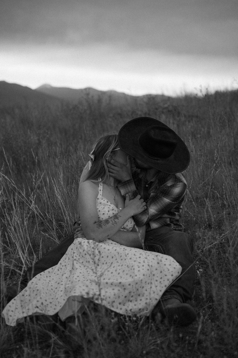 Black and white photo of a romantic couple embracing in a field, man in cowboy hat, mountains in background at dusk.