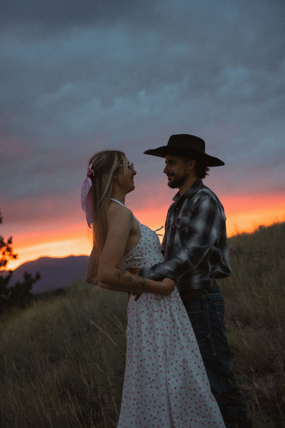 Romantic couple embracing in field at vivid sunset, man in cowboy hat and plaid shirt with woman in floral dress.