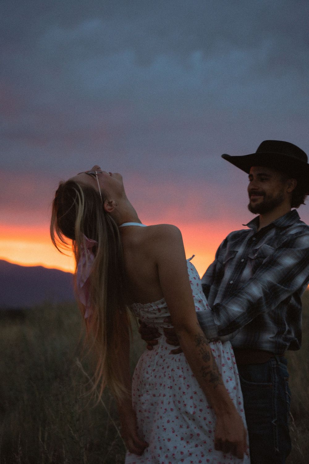 Couple dancing at sunset in a field, woman in floral dress with long hair, man in cowboy hat and plaid shirt.