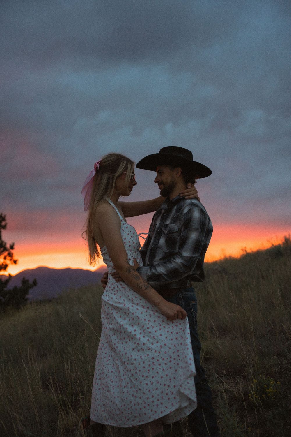 Couple embracing at sunset in a field, woman in floral dress and man in cowboy hat, with vibrant pink and orange sky.