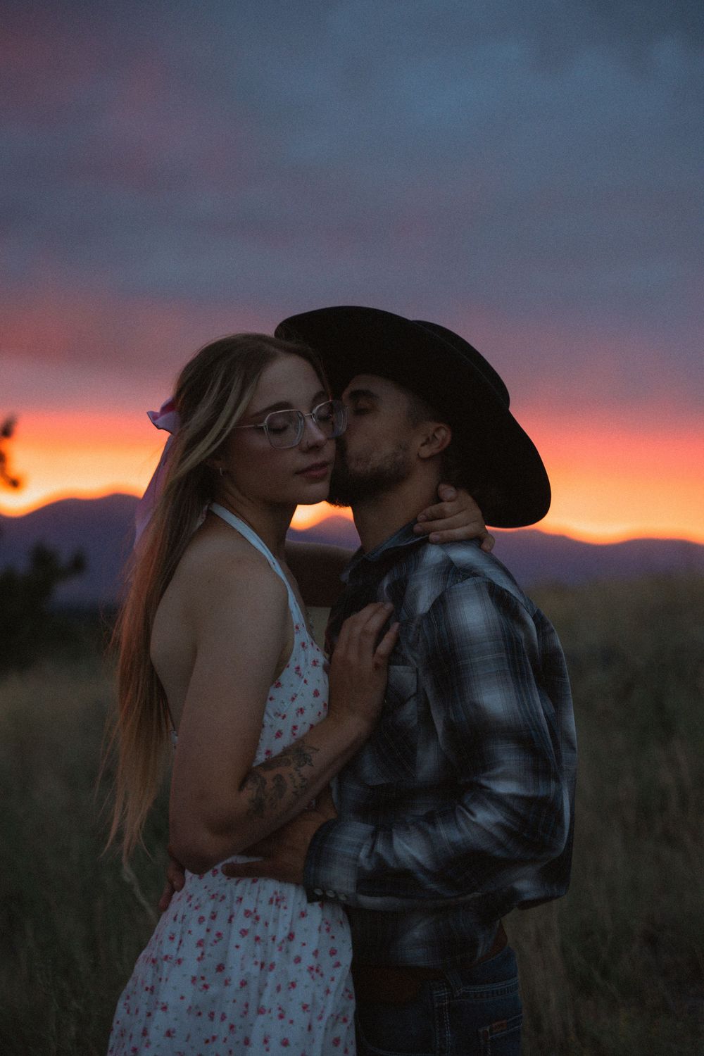 Couple embracing at sunset, man in cowboy hat kissing woman in floral dress, colorful orange sky backdrop.