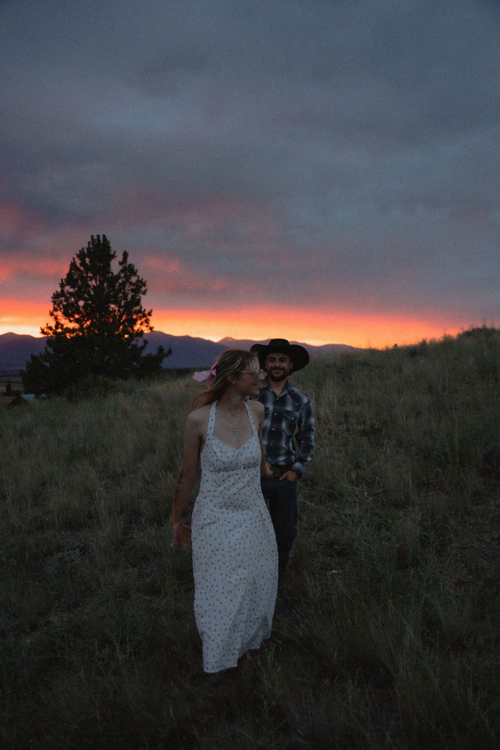 Couple walking through golden meadow grass at sunset with dramatic pink and purple sky and mountain backdrop.