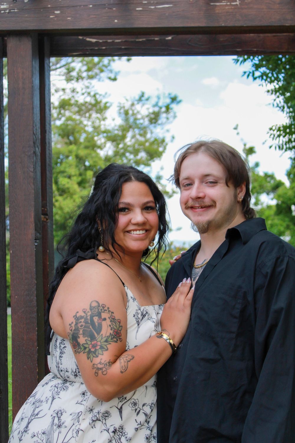A smiling couple poses outdoors under a wooden pergola, woman with tattoos in floral dress, man in black shirt.