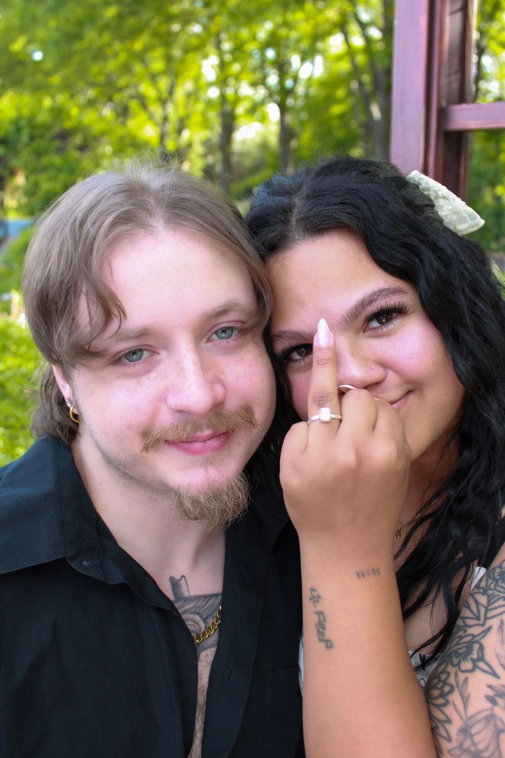A smiling couple poses outdoors, the woman showing off her engagement ring while leaning close to her partner.