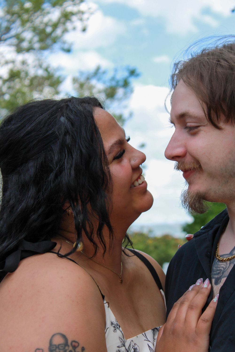 A smiling couple gazes lovingly at each other outdoors, woman with dark hair and tattoos, man with light hair and goatee.