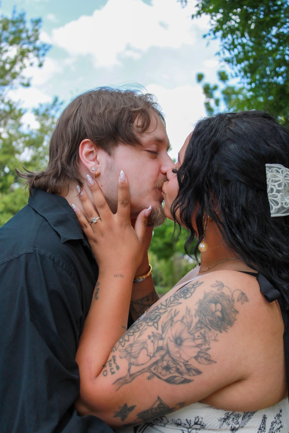 Couple sharing a romantic kiss outdoors, woman with floral tattoos and white hair accessory, man in black shirt.