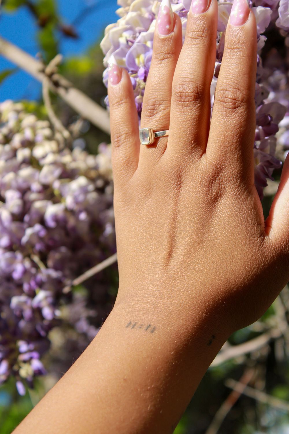 Close-up of woman's hand with square gemstone ring and small tattoo near purple wisteria flowers.