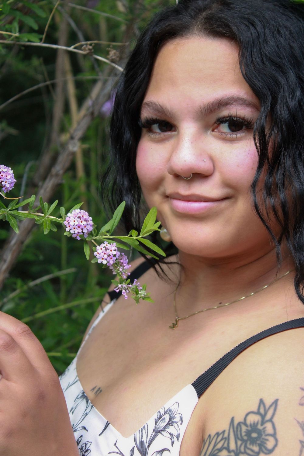 Young woman with dark curly hair smiles holding purple wildflowers, showing tattoos, wearing floral top outdoors.