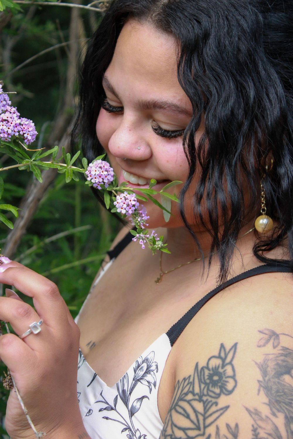 Smiling brunette woman with shoulder tattoo and pearl earring smelling purple wildflowers outdoors.