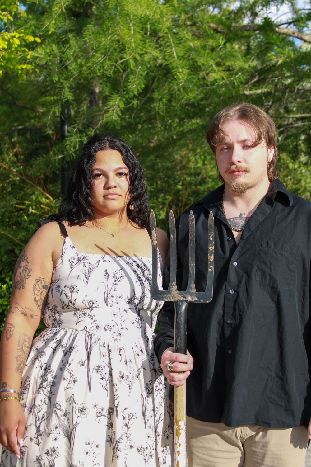 Couple posing seriously outdoors holding a pitchfork, woman in floral dress with tattoos, man in black shirt, green trees behind.