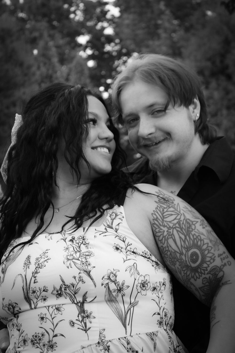 Black and white photo of a smiling couple embracing outdoors, man with tattoos and woman in floral dress.