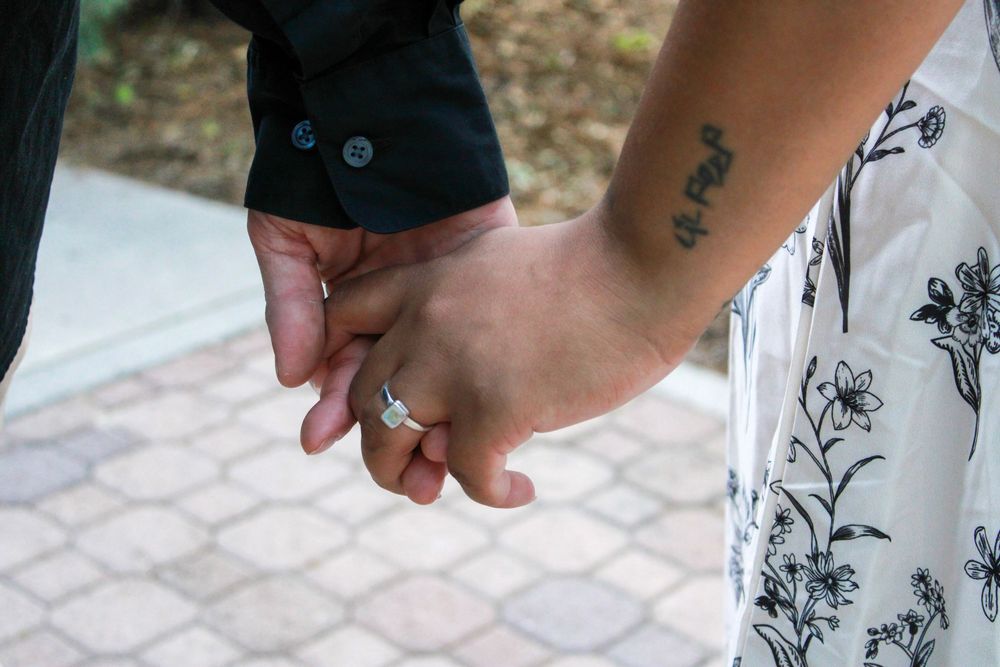 Close-up of a couple holding hands, woman wearing an engagement ring and floral dress with a wrist tattoo.