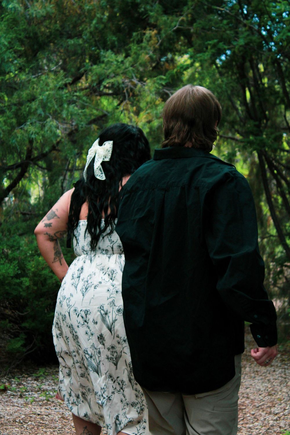 Couple walking hand in hand through a lush green forest path, woman in floral dress with white bow.