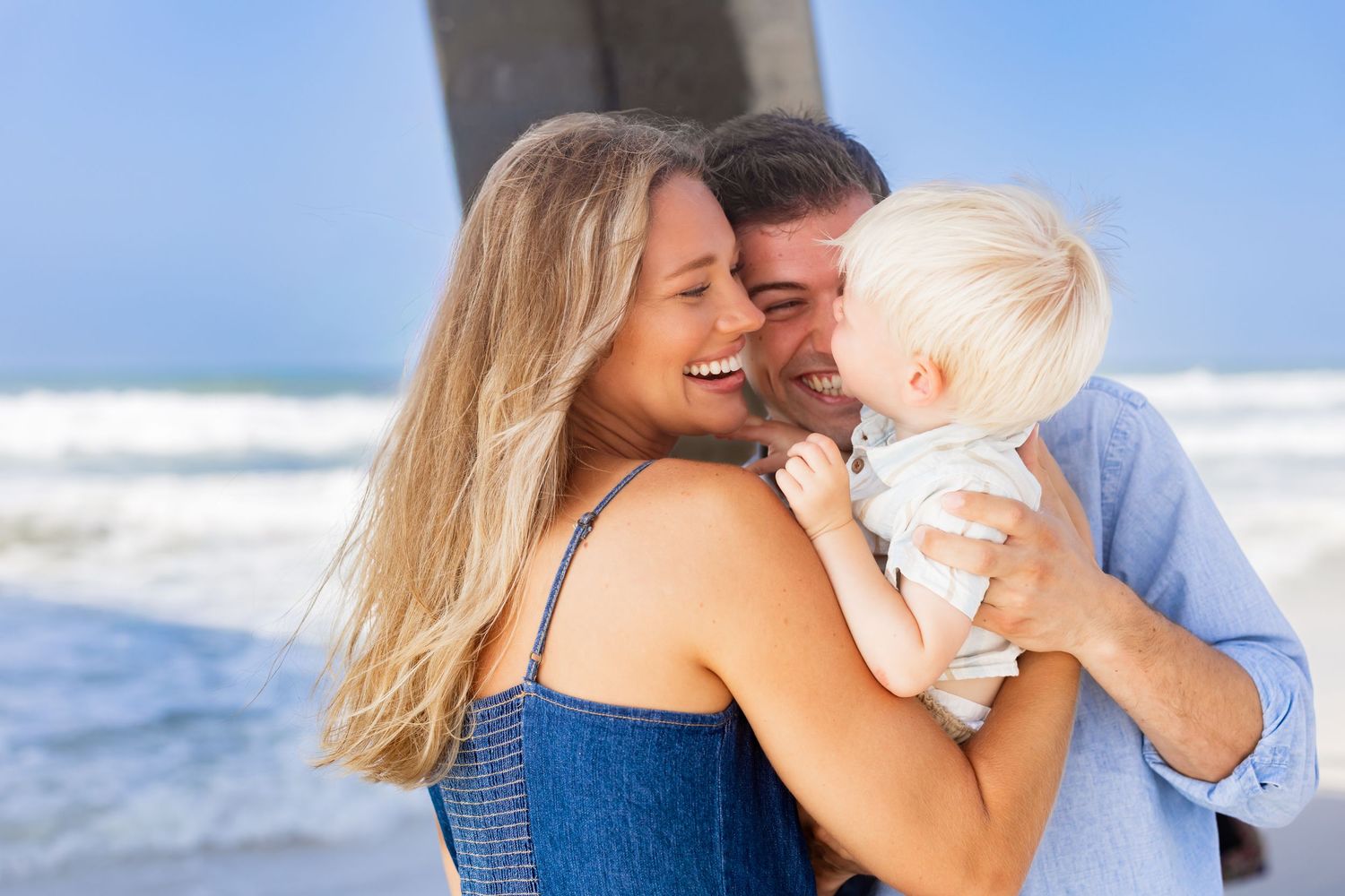 Happy young family with blonde baby sharing a joyful moment on a sunny beach near the ocean.