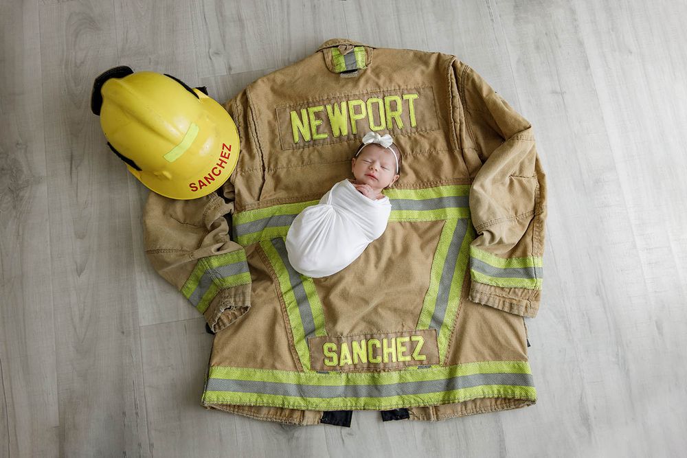 Newborn baby swaddled in white, lying on a Newport Sanchez firefighter jacket with yellow helmet beside it.