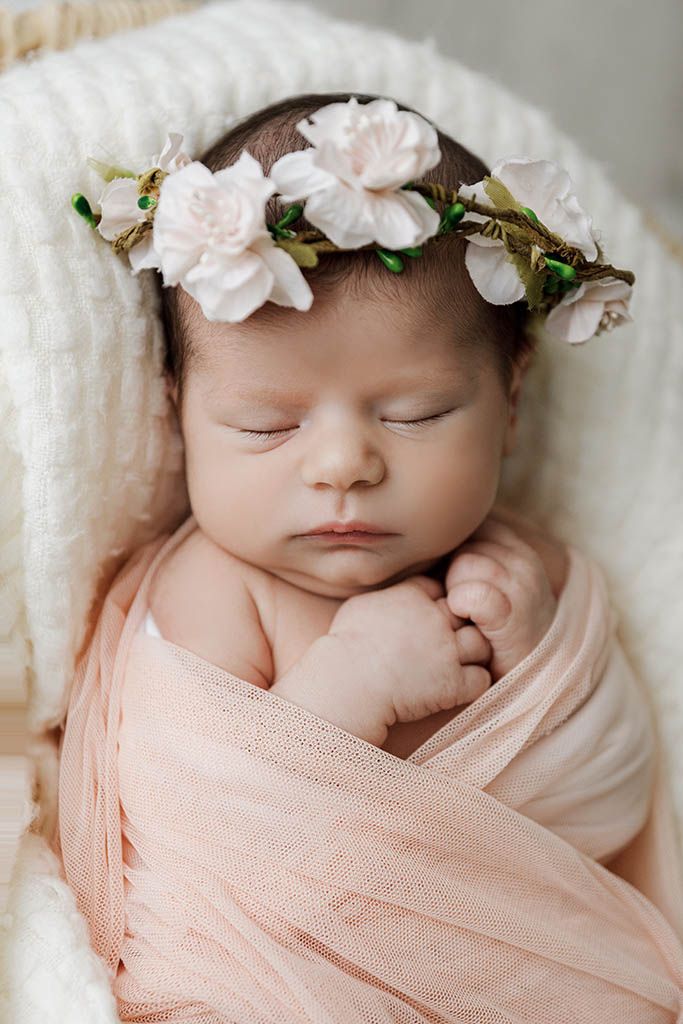 Sleeping newborn baby wrapped in peach fabric wearing a delicate white flower crown in a cozy basket.