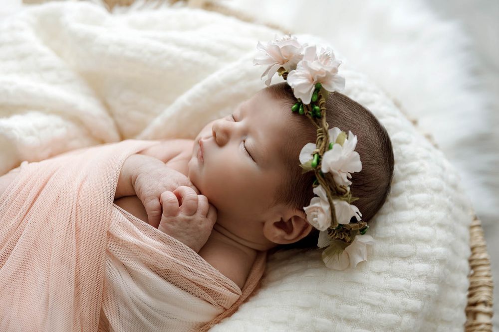 Sleeping newborn baby wrapped in peach swaddle wearing a delicate white floral crown on a soft white blanket.