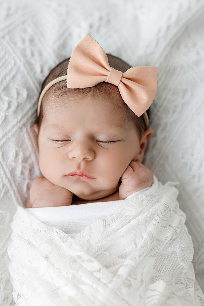 Sleeping newborn baby girl swaddled in white lace wrap wearing a peach bow headband on a white textured background.