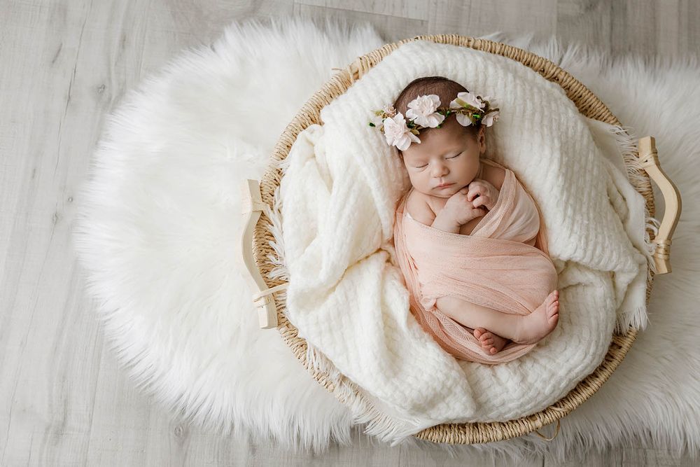 Newborn baby girl wrapped in peach swaddle sleeping in wicker basket with floral crown on white fur rug.