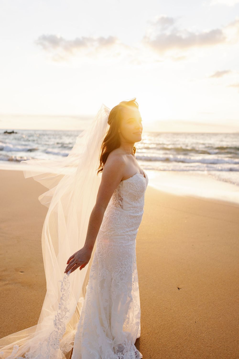 Bride in lace wedding gown and flowing veil stands on sandy beach at golden sunset with ocean waves behind her.
