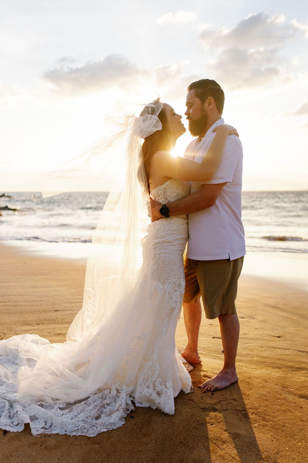 Bride and groom embrace on a sandy beach at golden sunset, her lace gown and veil flowing in the breeze.
