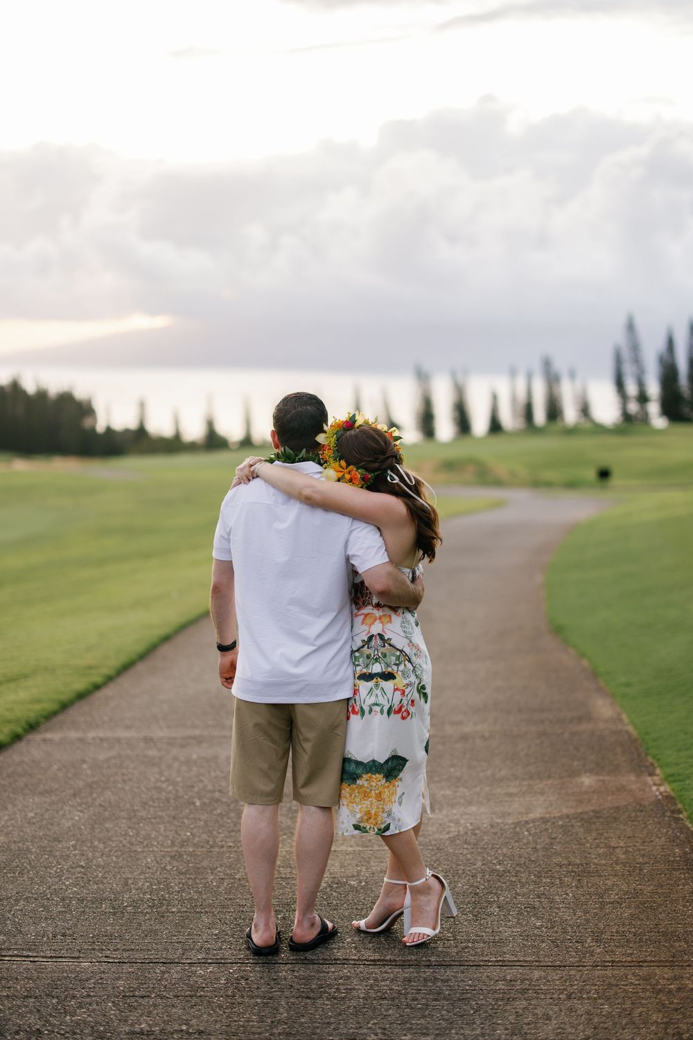 Couple embracing on a path in Hawaii, woman wearing floral dress and flower crown, lush green golf course backdrop.