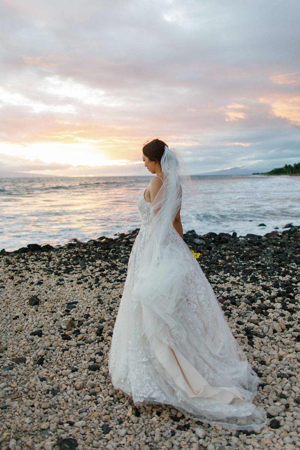 Bride in lace wedding gown and veil stands on rocky beach gazing at sunset over ocean waves.