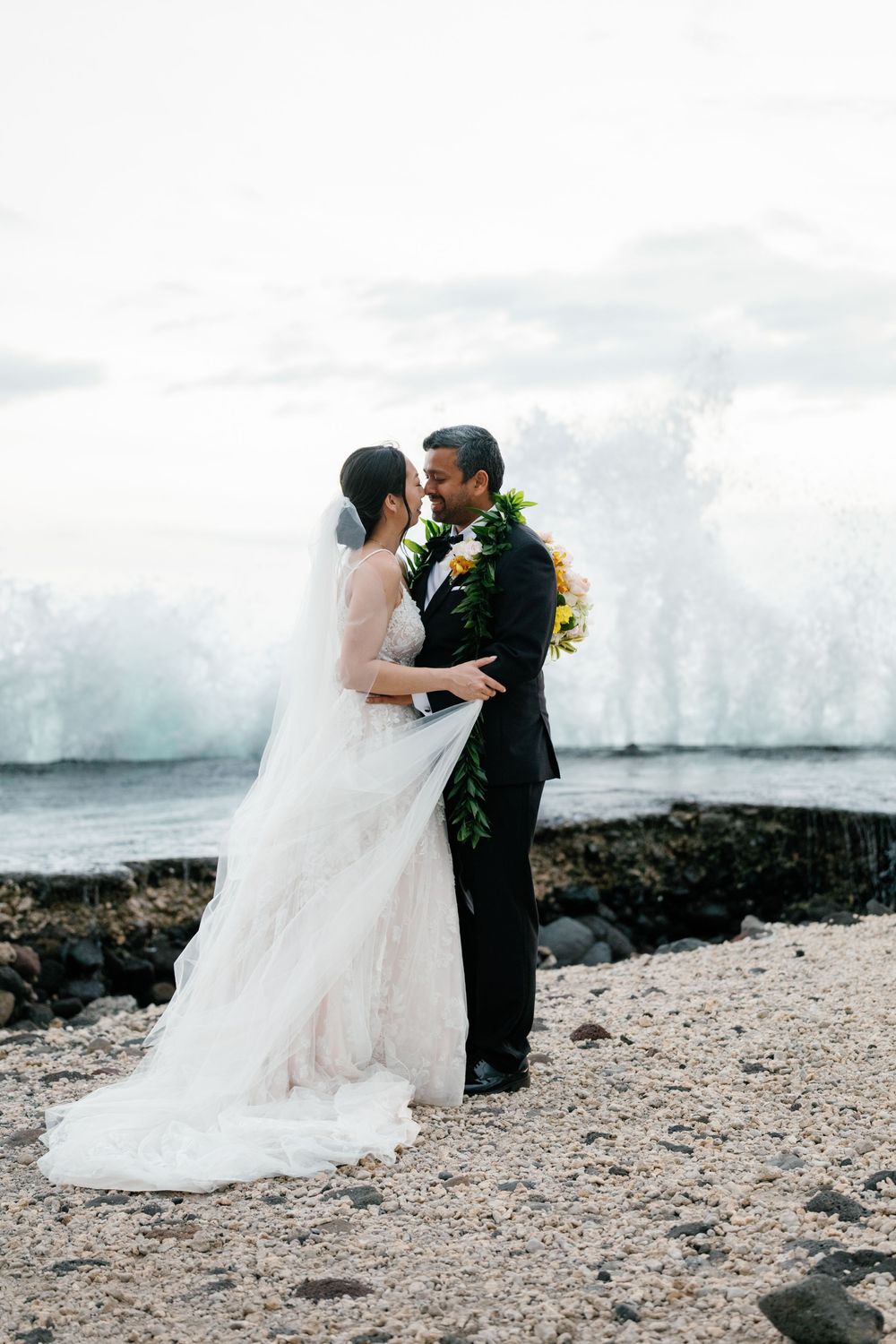 Bride and groom share a romantic kiss on a rocky Hawaiian beach with dramatic ocean waves crashing behind them.