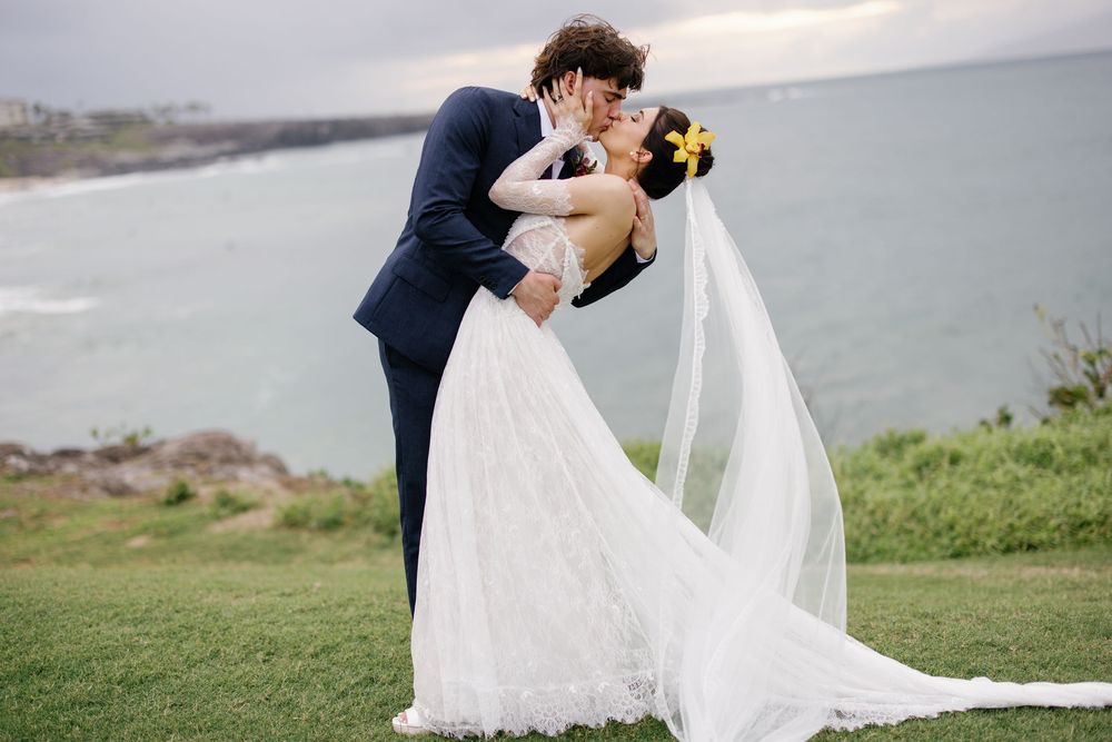 Bride and groom share a romantic kiss at an oceanside wedding, with a flowing white gown and veil in a coastal setting.