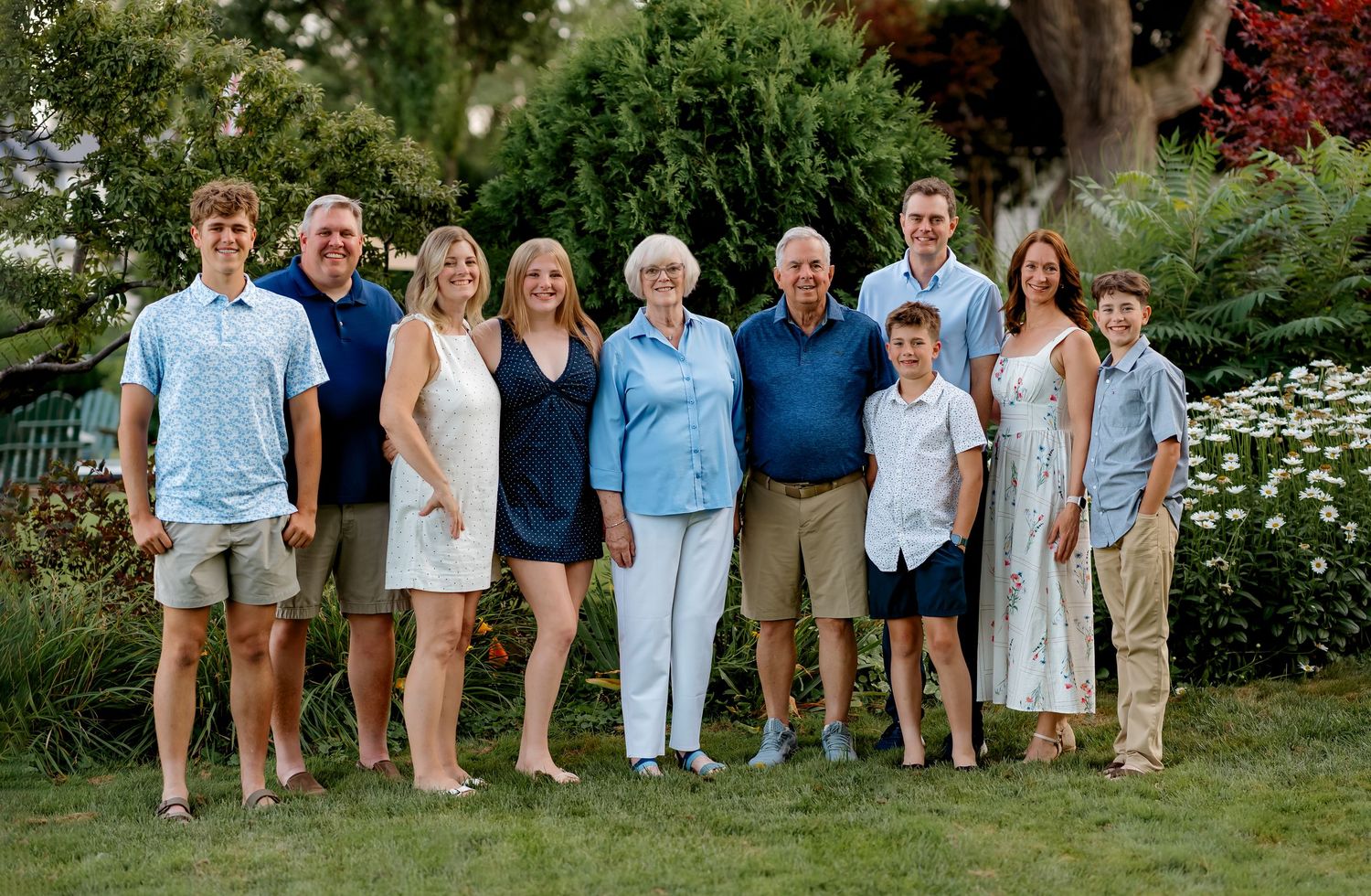 Large family group portrait outdoors on green grass with trees, smiling adults and children in coordinated blue and neutral outfits.