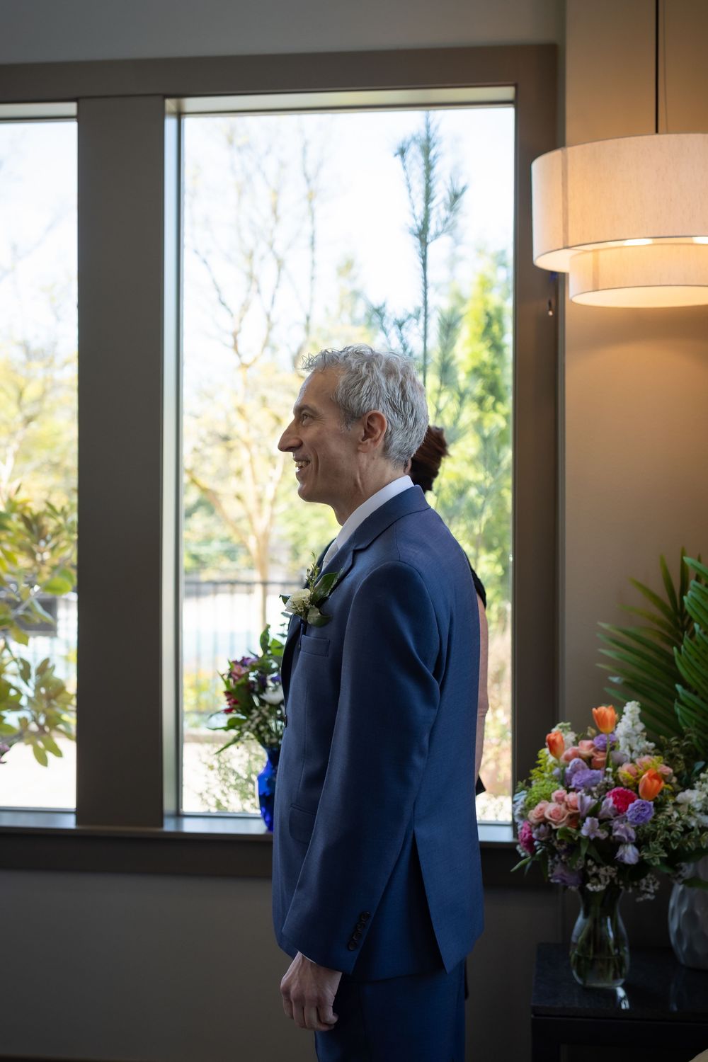 Smiling groom in blue suit with boutonniere stands by window in bright indoor wedding venue.