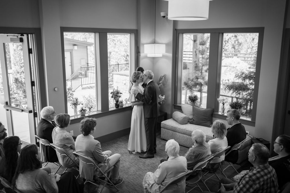 Black and white photo of a couple exchanging vows at an intimate indoor wedding ceremony with seated guests.