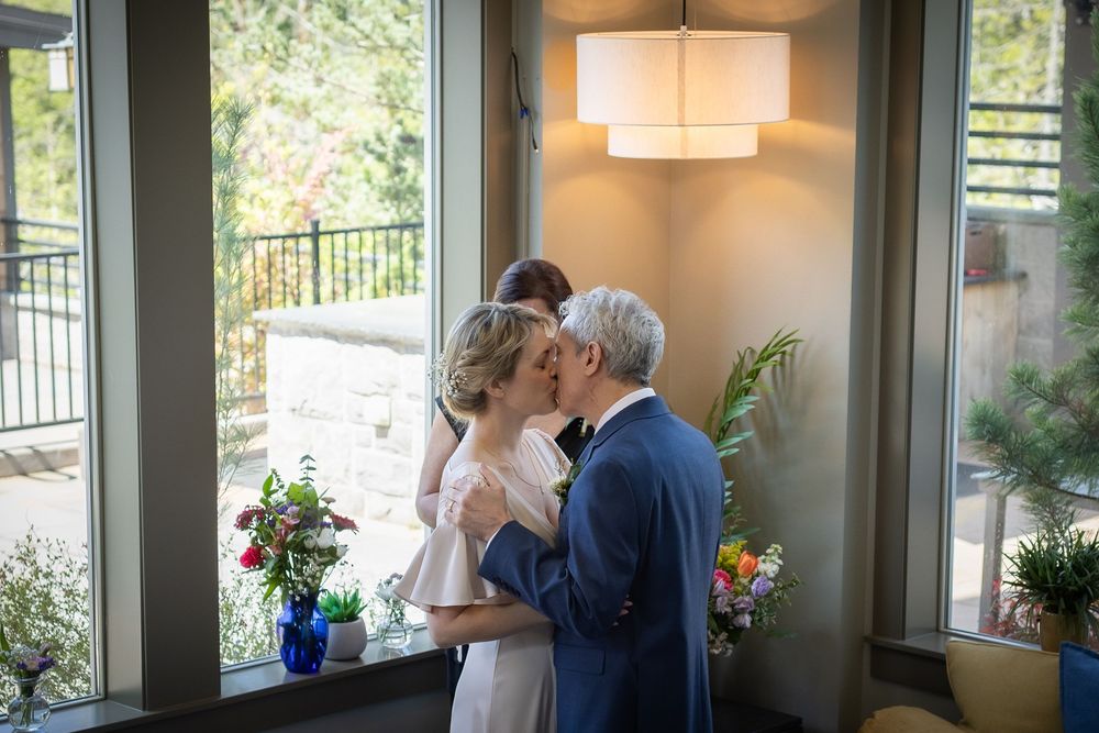 Couple sharing a first kiss at indoor wedding ceremony with floral arrangements and large windows.