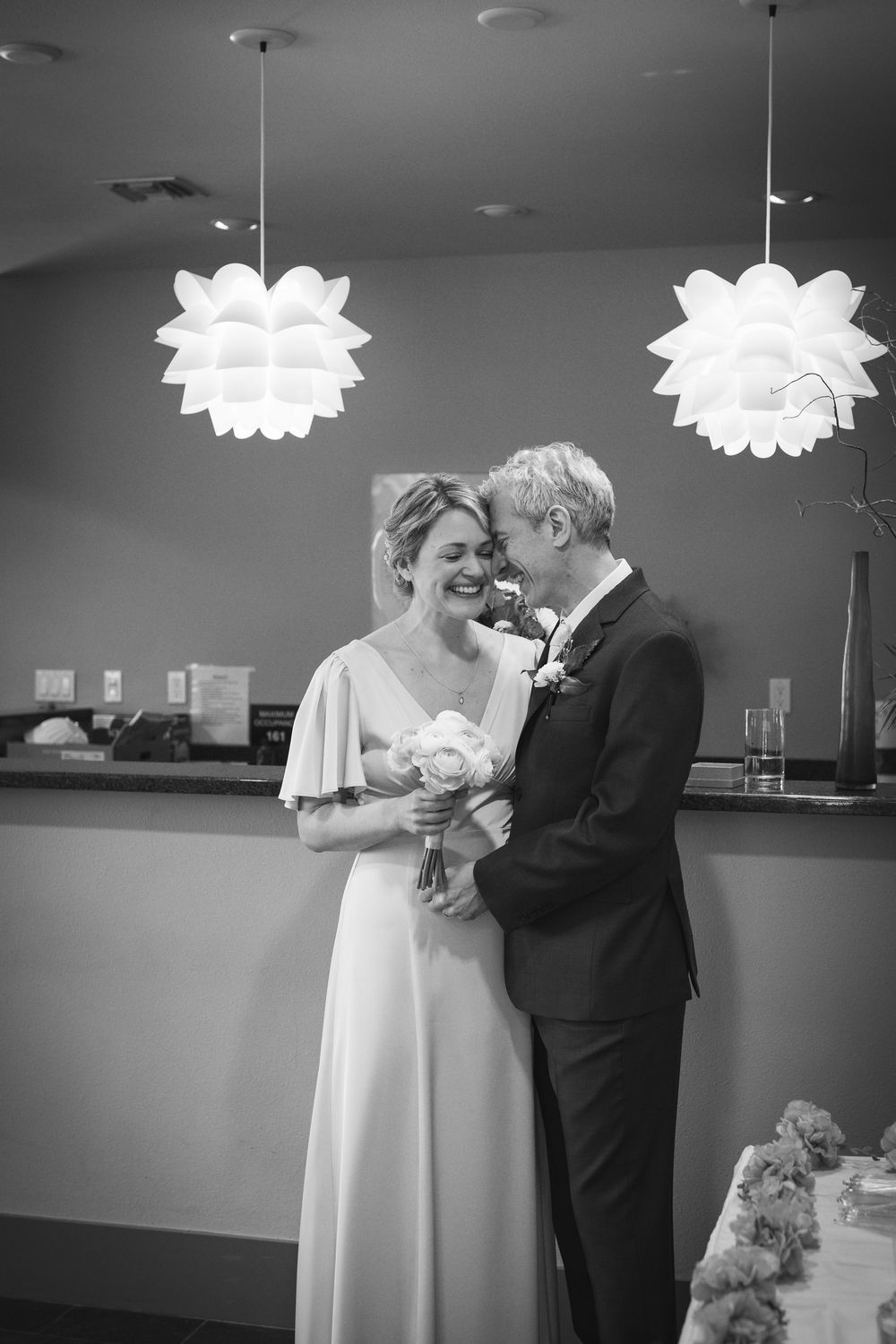 Black and white photo of a smiling bride holding bouquet and groom sharing a joyful moment under floral pendant lights.