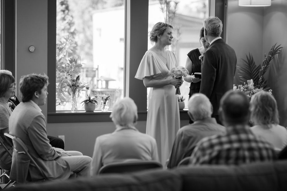 Black and white photo of a wedding ceremony with guests seated watching a couple exchange vows indoors.