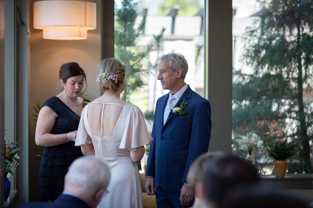Elegant indoor wedding ceremony with bride in white dress, groom in blue suit, and officiant in black dress exchanging vows.