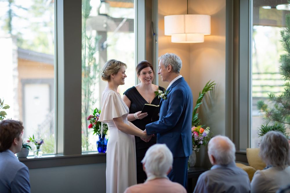 Couple exchanging vows at indoor wedding ceremony with officiant, guests seated, floral arrangements, and large windows in background.
