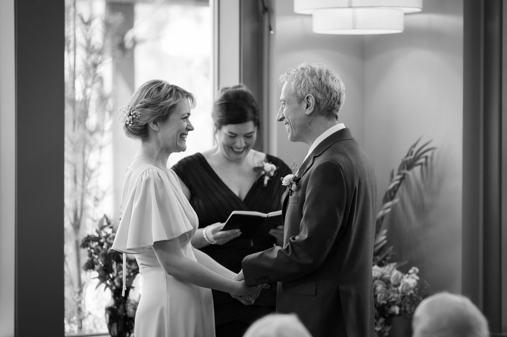 Black and white photo of a bride and groom exchanging vows with a female officiant during an intimate wedding ceremony.